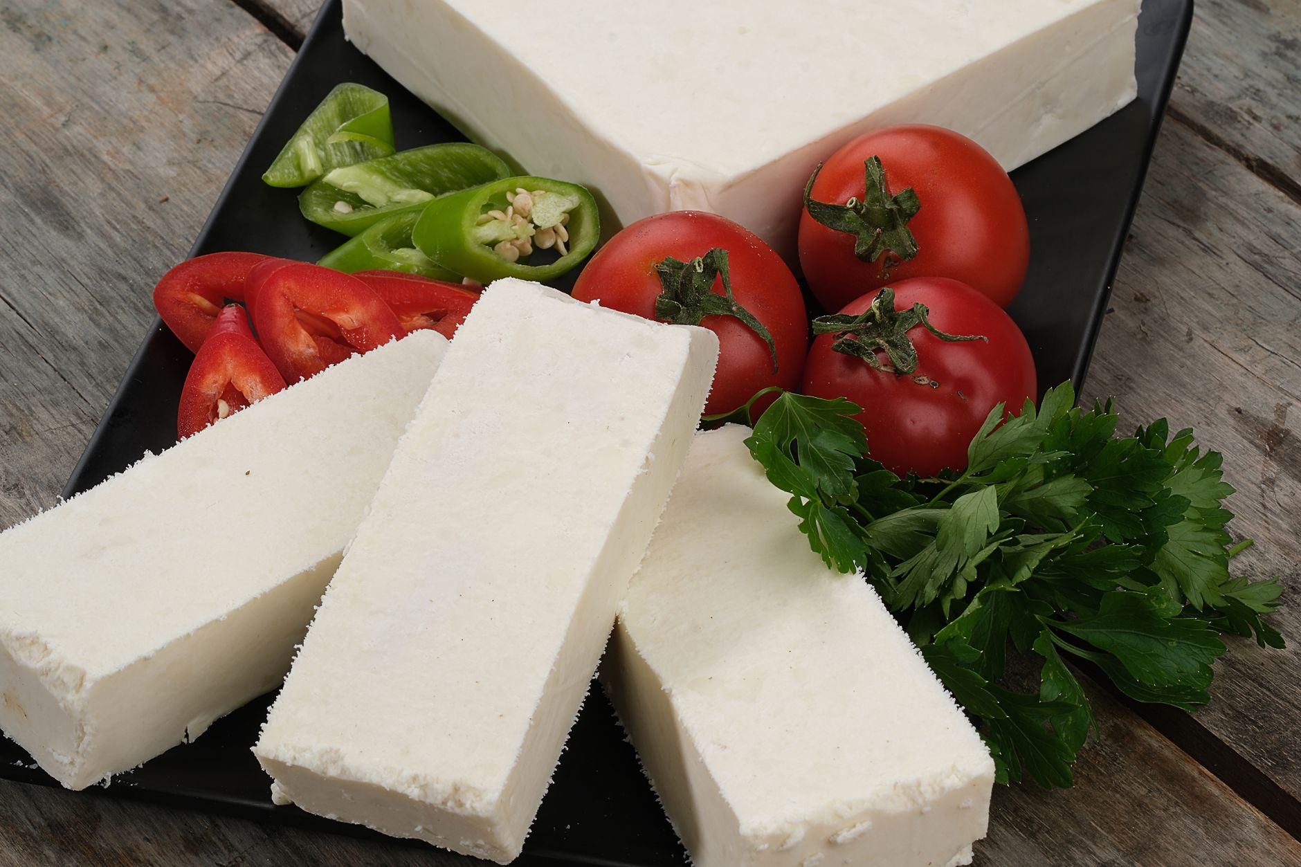 Tres bloques alargados de queso blanco firme sobre una tabla negra, con tomates, pimientos y perejil