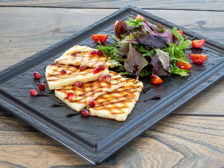 Cuatro lonchas de queso halloumi con marcas de parrilla sobre una pizarra negra, con ensalada y granada