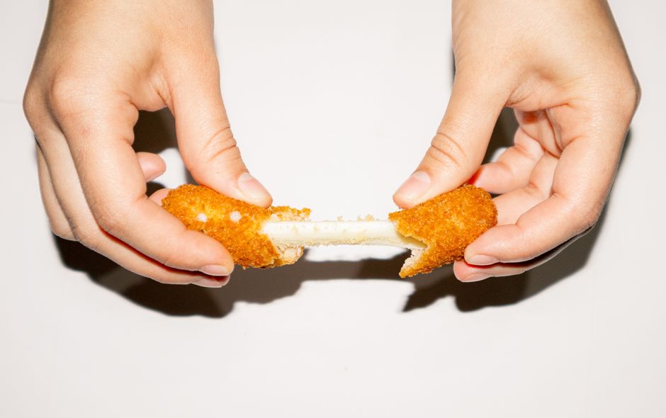 Dos manos separan un palito de queso empanizado y frito, del que se estira un hilo de queso blanco