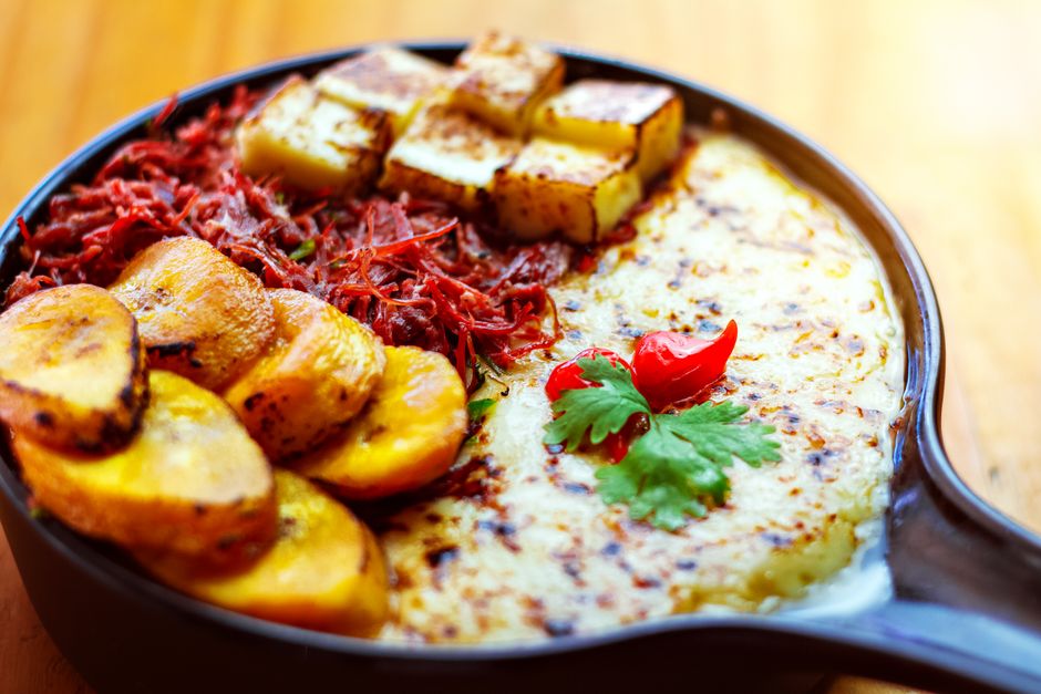 Sartén de hierro con rodajas de plátano frito, cubos de queso dorado, carne desmenuzada y queso gratinado