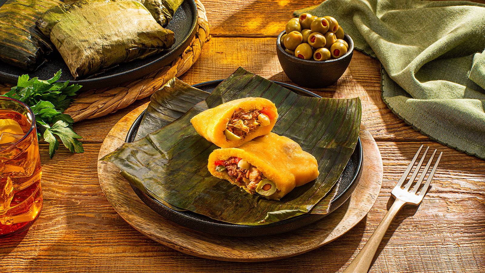Plato de tamales envueltos en hojas de plátano, uno abierto con relleno, junto a un tazón de aceitunas