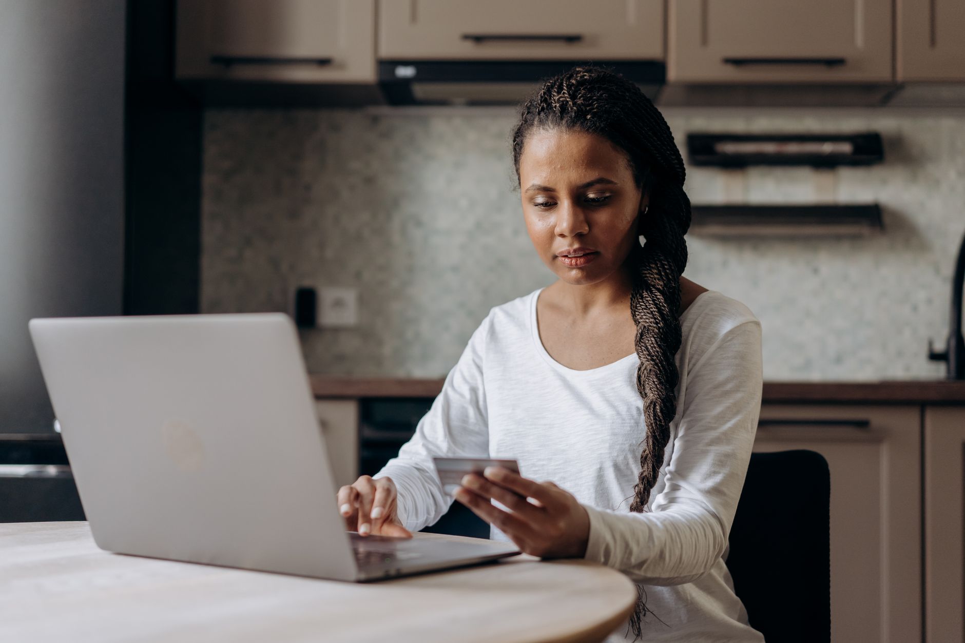 Mujer joven sentada a la mesa de su cocina con la laptop abierta y su tarjeta bancaria en la mano para pagar un trámite en línea