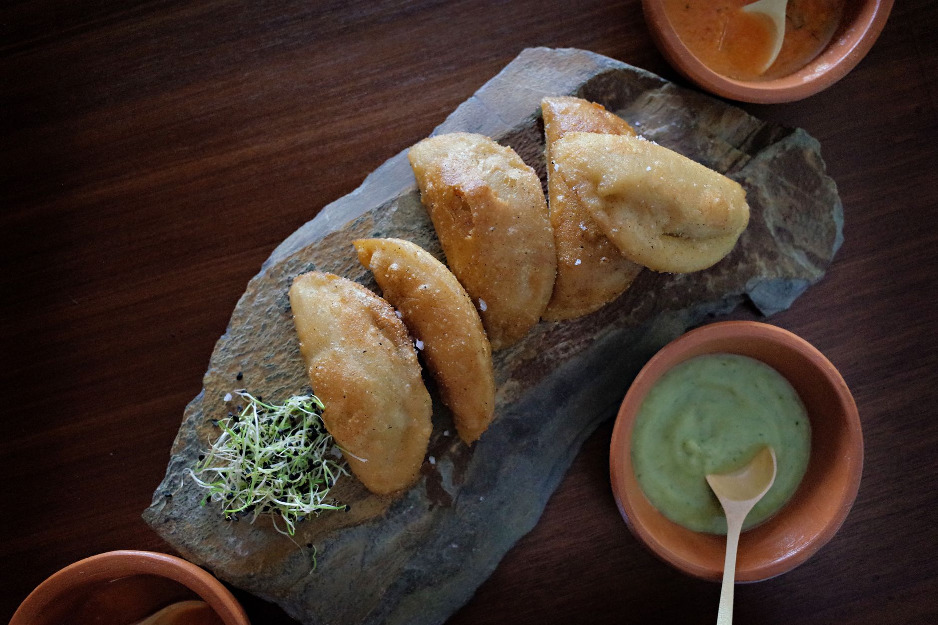 Empanadas fritas servidas sobre una piedra con dos cuencos de barro, uno de salsa verde cremosa de aguacate y otro de salsa roja picante