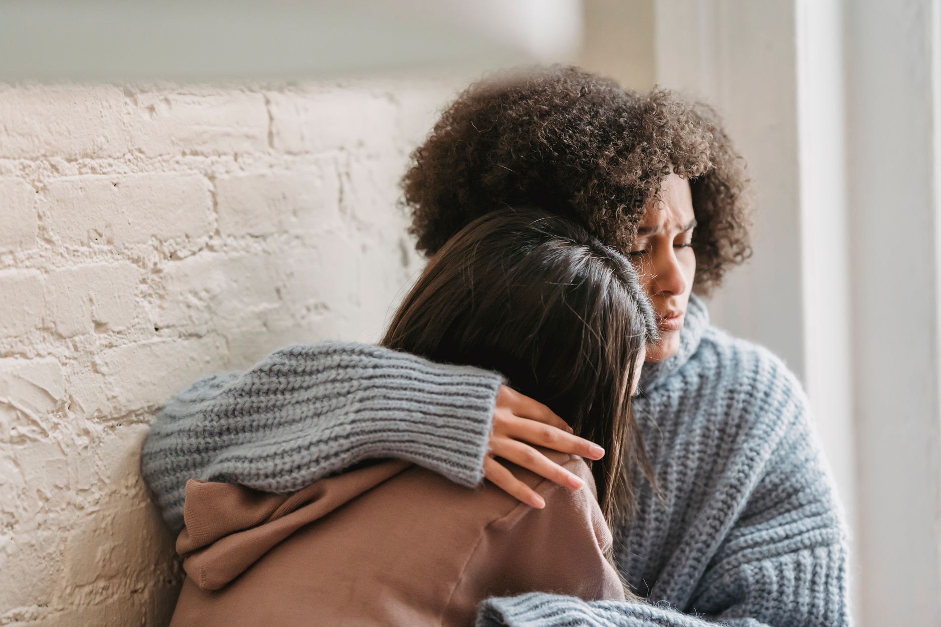 Una mujer abraza y consuela a una amiga sentadas contra la pared de casa
