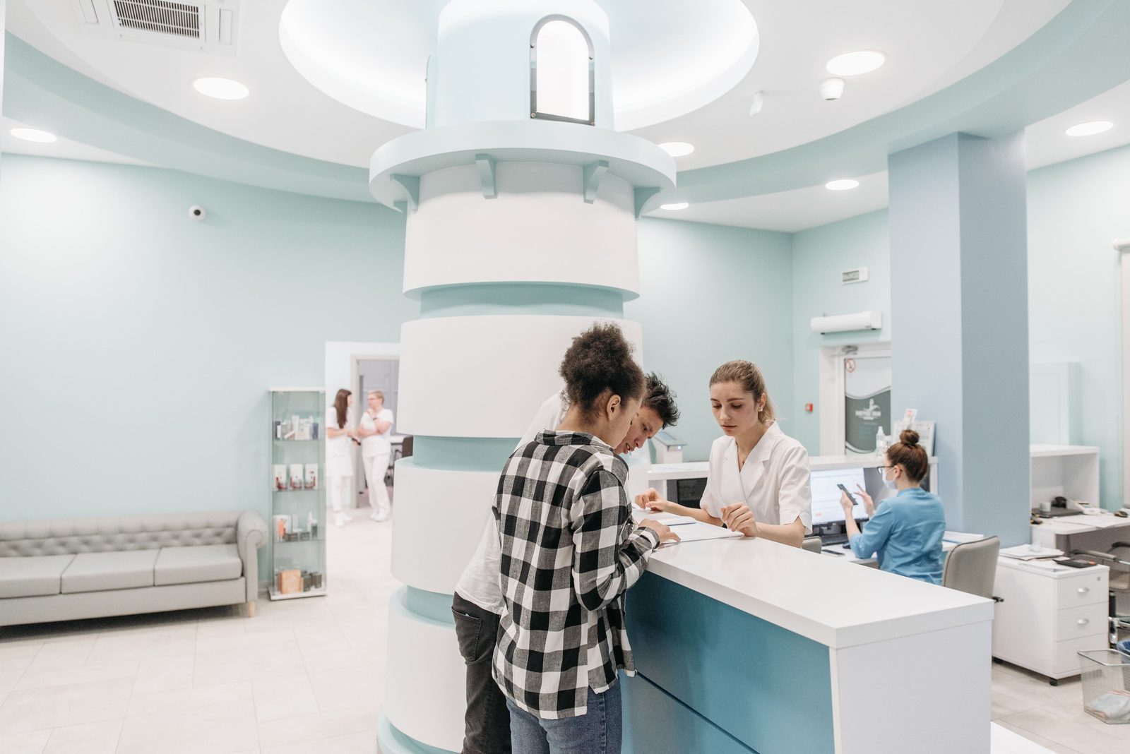 Una pareja joven llena un formulario apoyada en el mostrador de recepción de una clínica mientras una trabajadora de salud con bata blanca les señala el papel y les explica los pasos