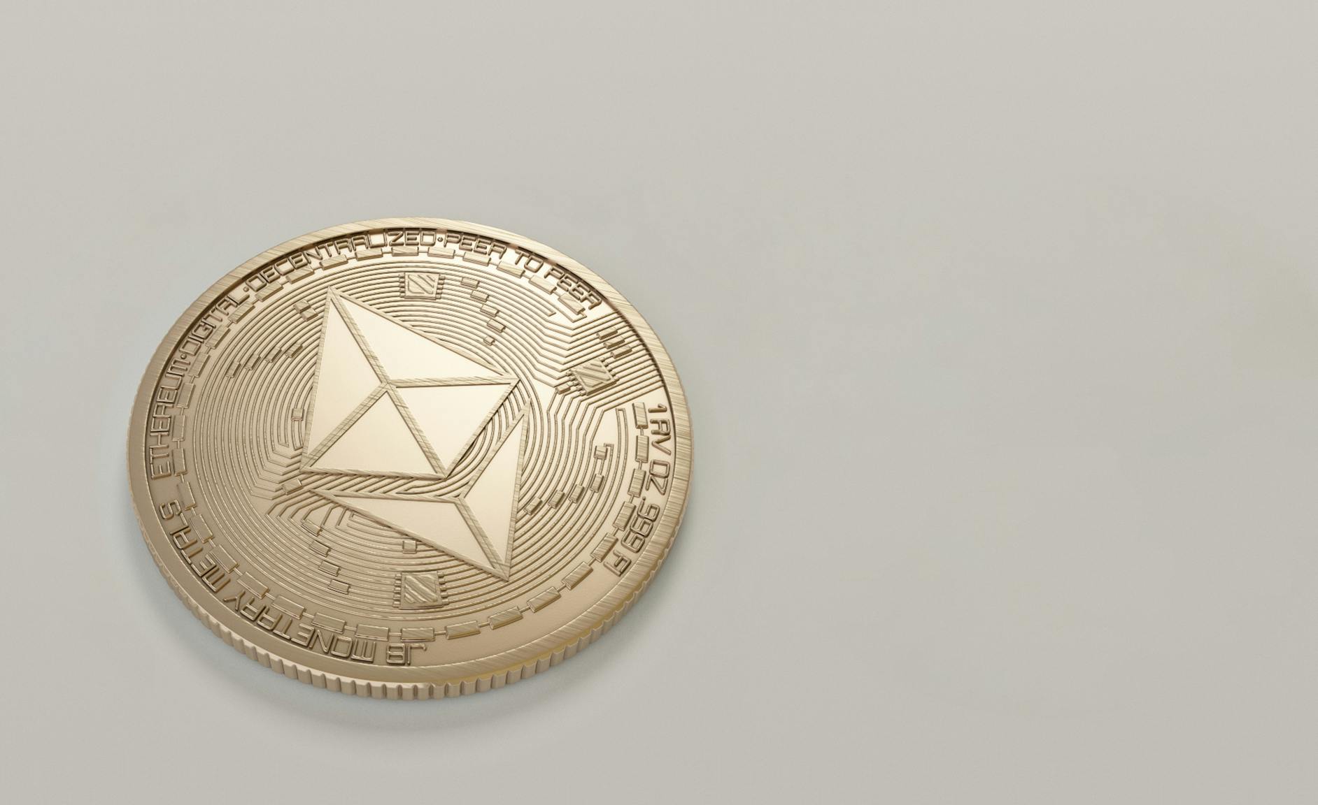 Moneda dorada de Ethereum con su logotipo hexagonal grabado y texto, sobre un fondo gris claro liso