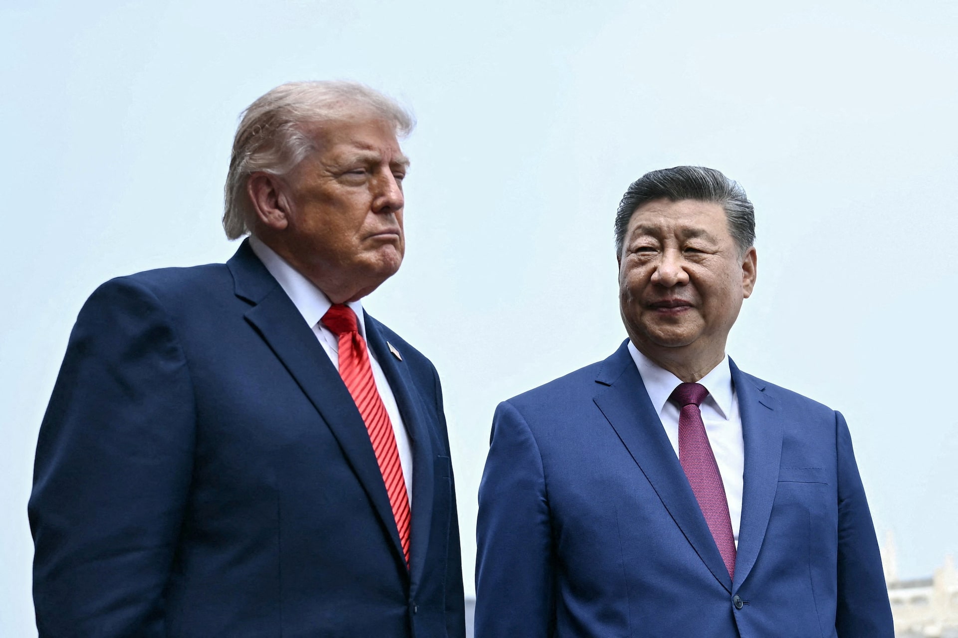 Donald Trump y Xi Jinping de pie con trajes y corbatas, mirando en direcciones opuestas bajo un cielo claro